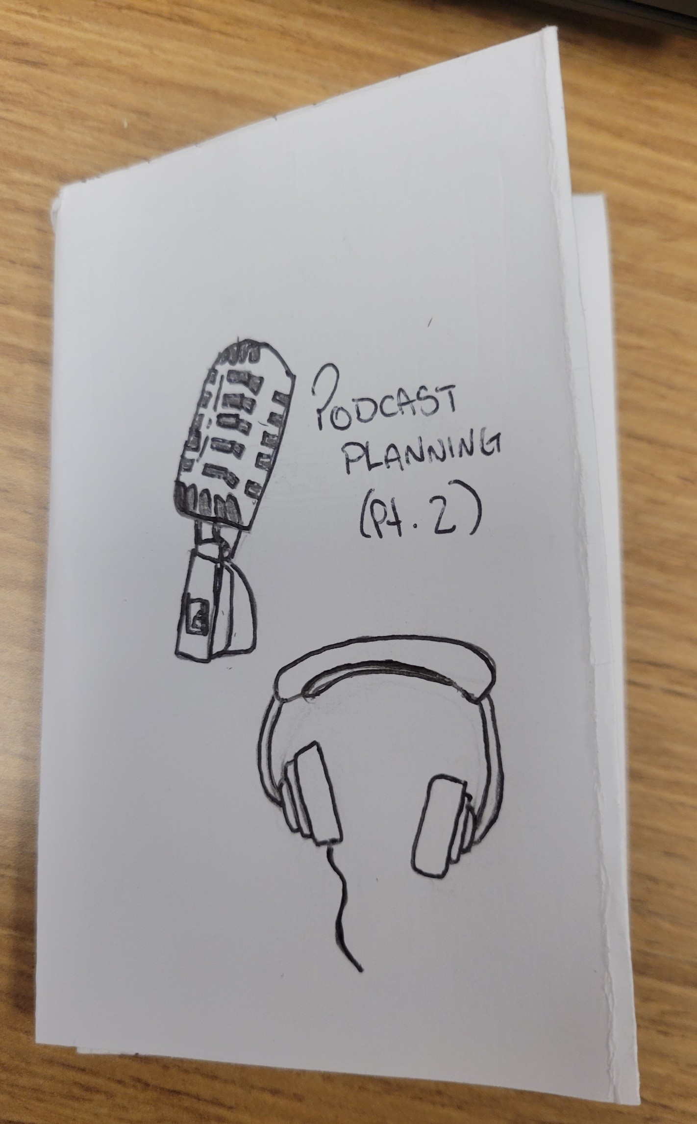 A black-and-white ink drawing on white paper featuring a vintage-style microphone on a stand and a pair of over-ear headphones. Handwritten text to the right of the microphone reads "PODCAST PLANNING (Pt. 2)". The drawing is placed on a light-colored wooden surface. Alt text generated by Google Gemini.