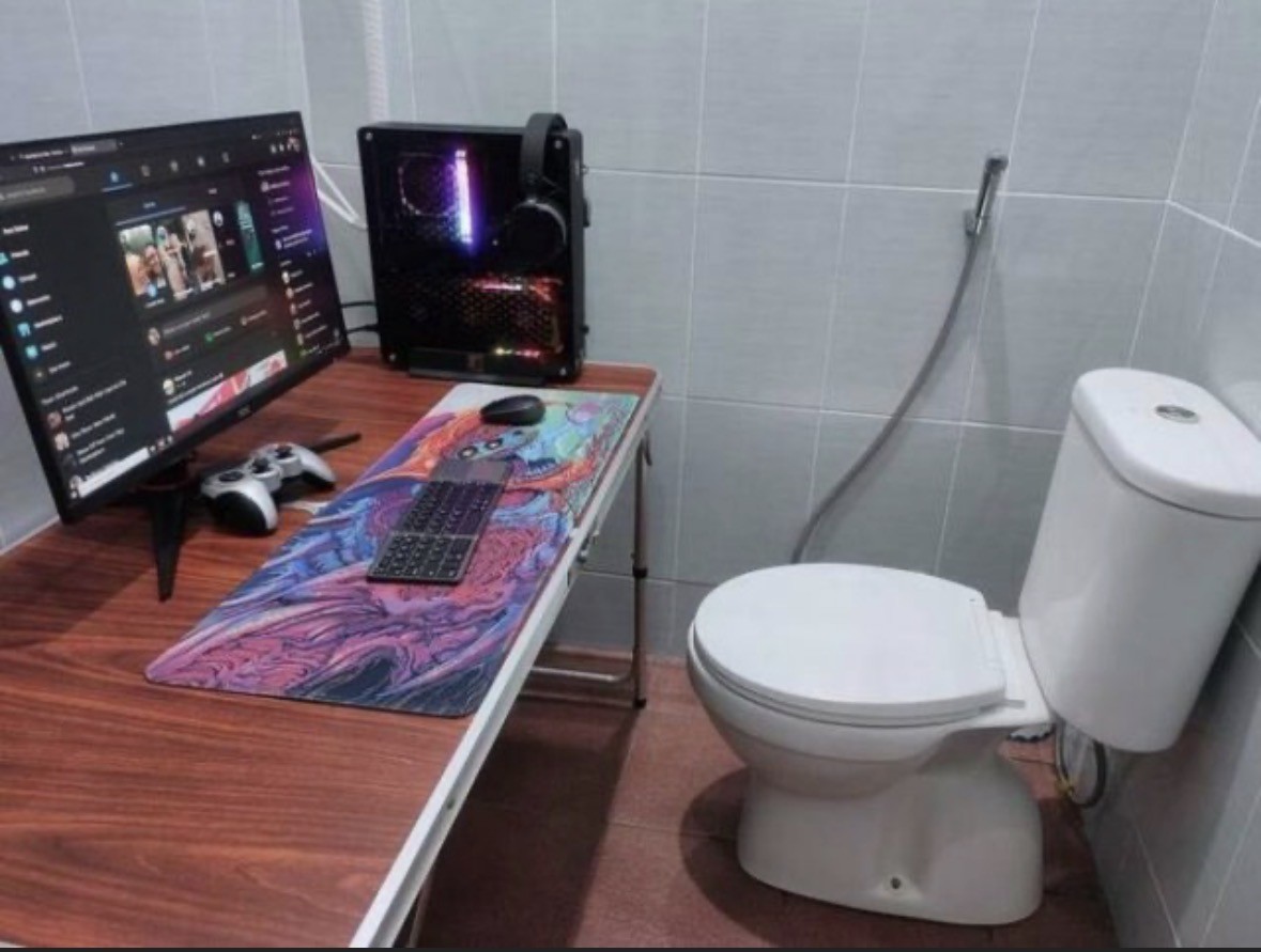 The room has a computer setup on the right side of the screen, complete with a desk of brown wood like material which has  a silver edge on it. On top of it is a black desktop computer and a monitor with text and apps on it. There’s a game controller under the monitor. It’s grey with black buttons. In front of that is a keyboard on a large mouse pad with a computer mouse on top of it. The chair for the desk is a white porcelain toilet. There’s a hose hanging on the wall beside it the walls are a white colour.