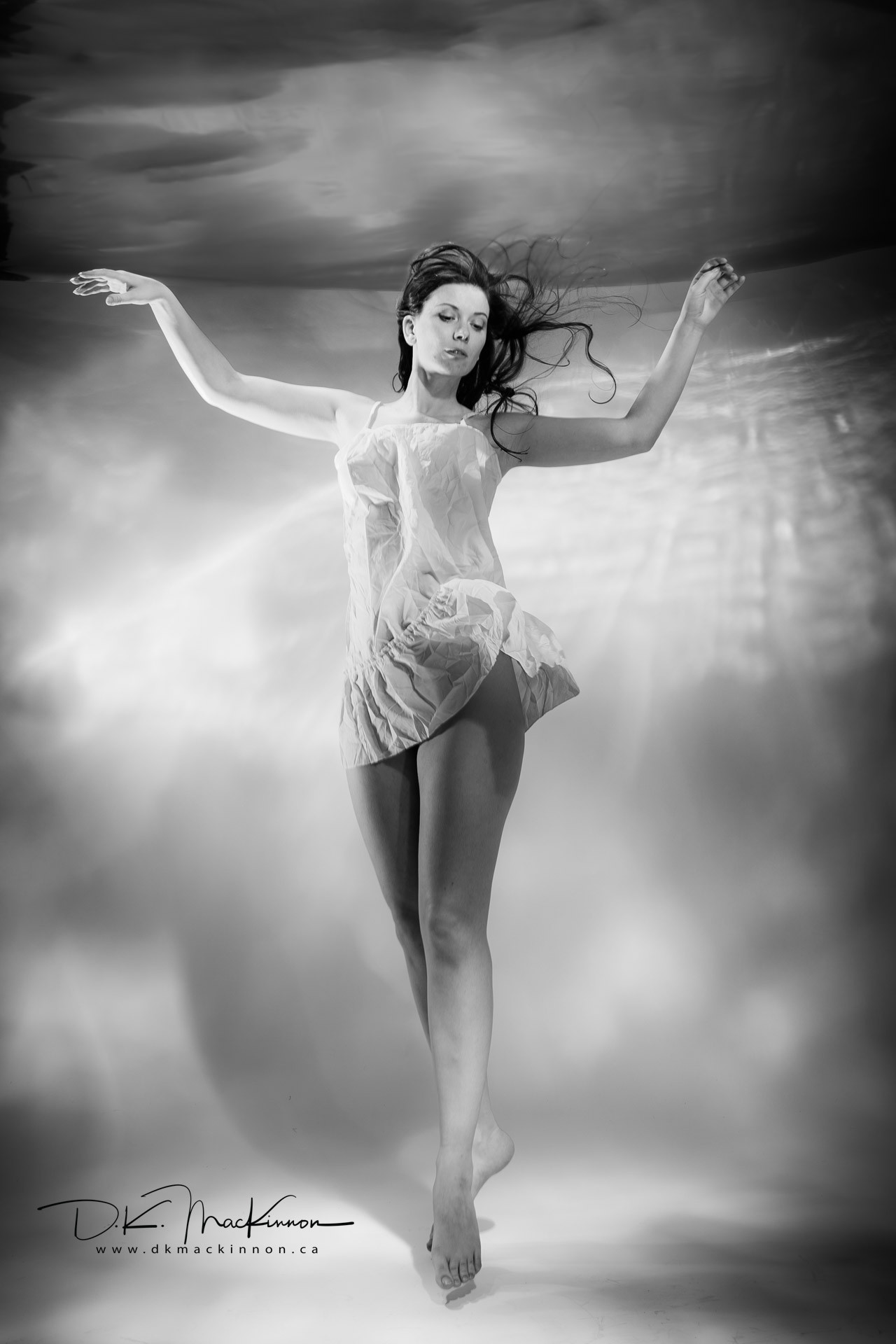 A black and white, full-body portrait of a woman wearing a sheer, ruffled dress, floating in water with her arms outstretched and hair flowing.