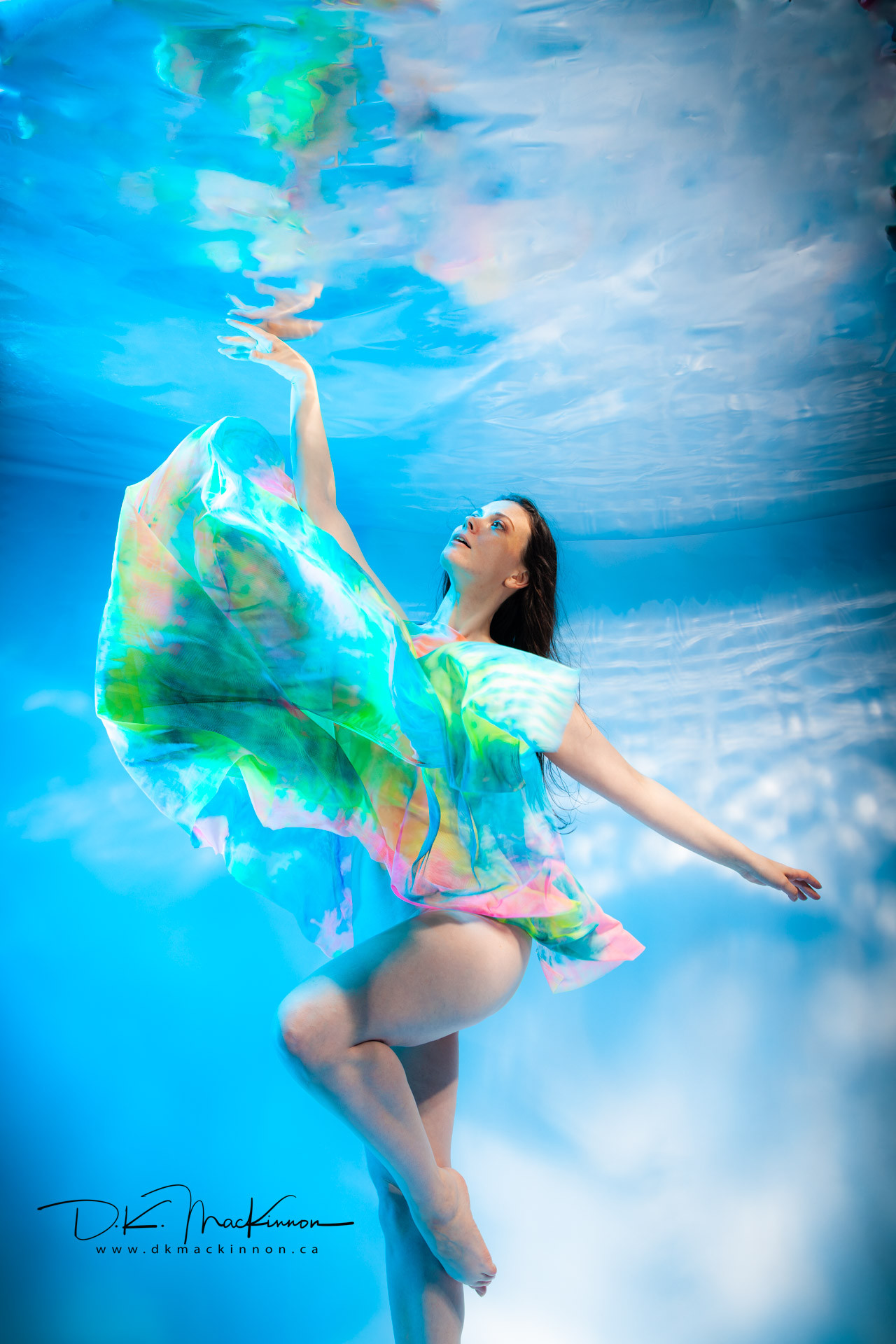 An underwater portrait of a woman in a brightly colored, flowing garment, with her arms outstretched and looking upwards towards the water's surface.